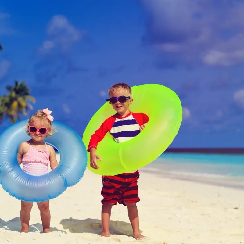 Two children with inflatable rings on a sandy beach, wearing sunglasses.