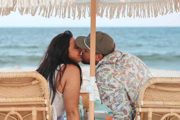 Couple kissing under a fringed umbrella on a beach.