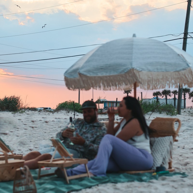 Two people relaxing under a beach umbrella at sunset on a sandy beach.