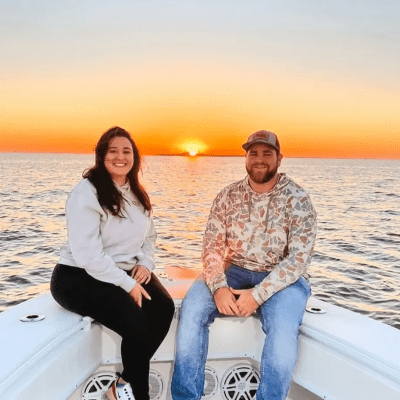 Two people sit on a boat's bow at sunset, smiling at the camera.