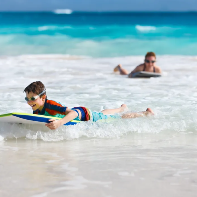 Child with goggles bodyboarding on waves, adult in background.